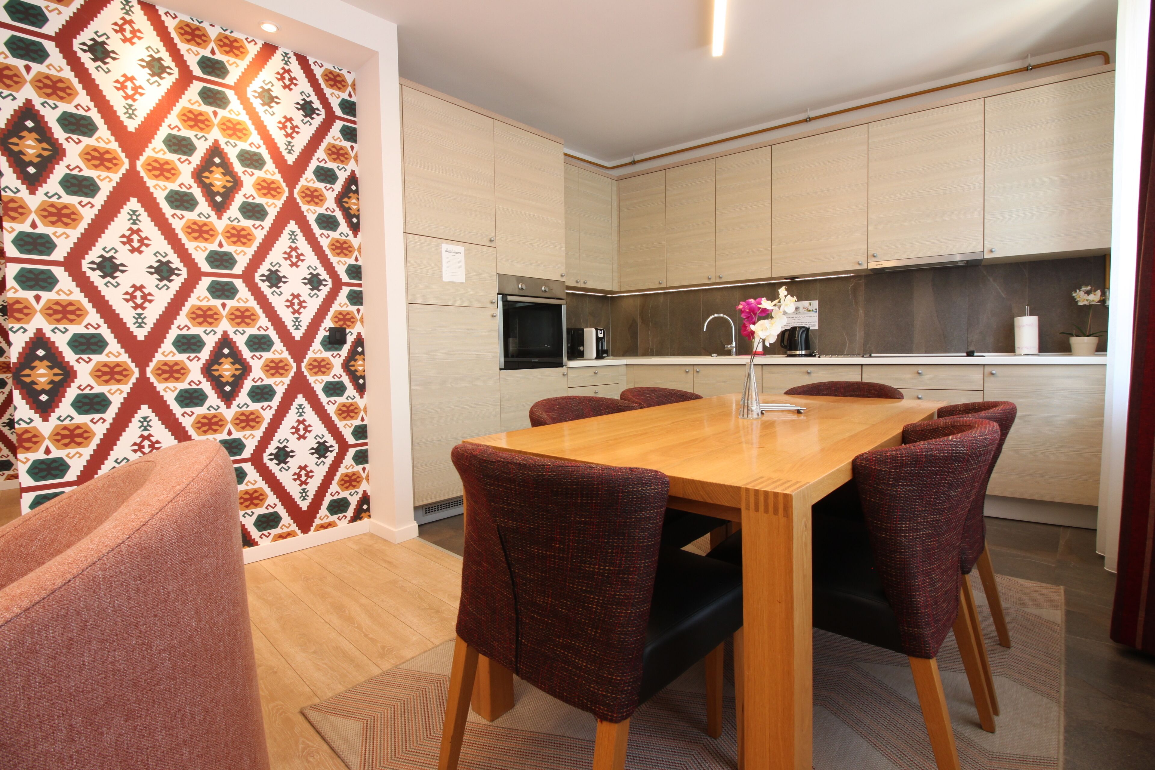 A modern kitchen with light wood cabinets, a central island, and a dining area with a wooden table and red upholstered chairs.