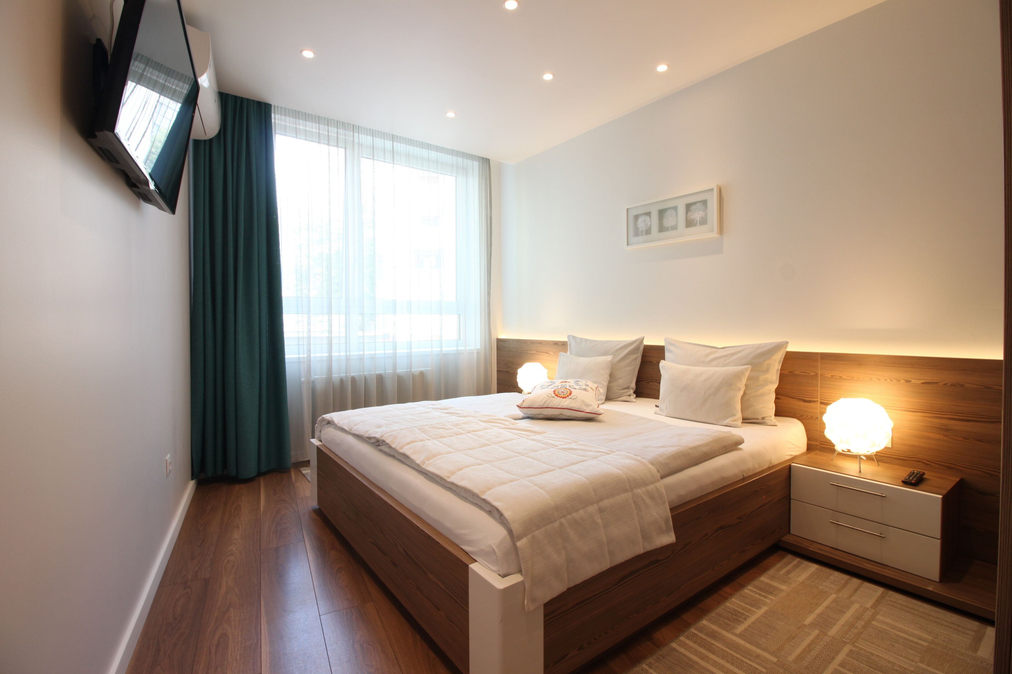 Une chambre à coucher moderne avec un lit en bois, des lampes de chevet, un téléviseur fixé au mur et une grande fenêtre avec des rideaux.