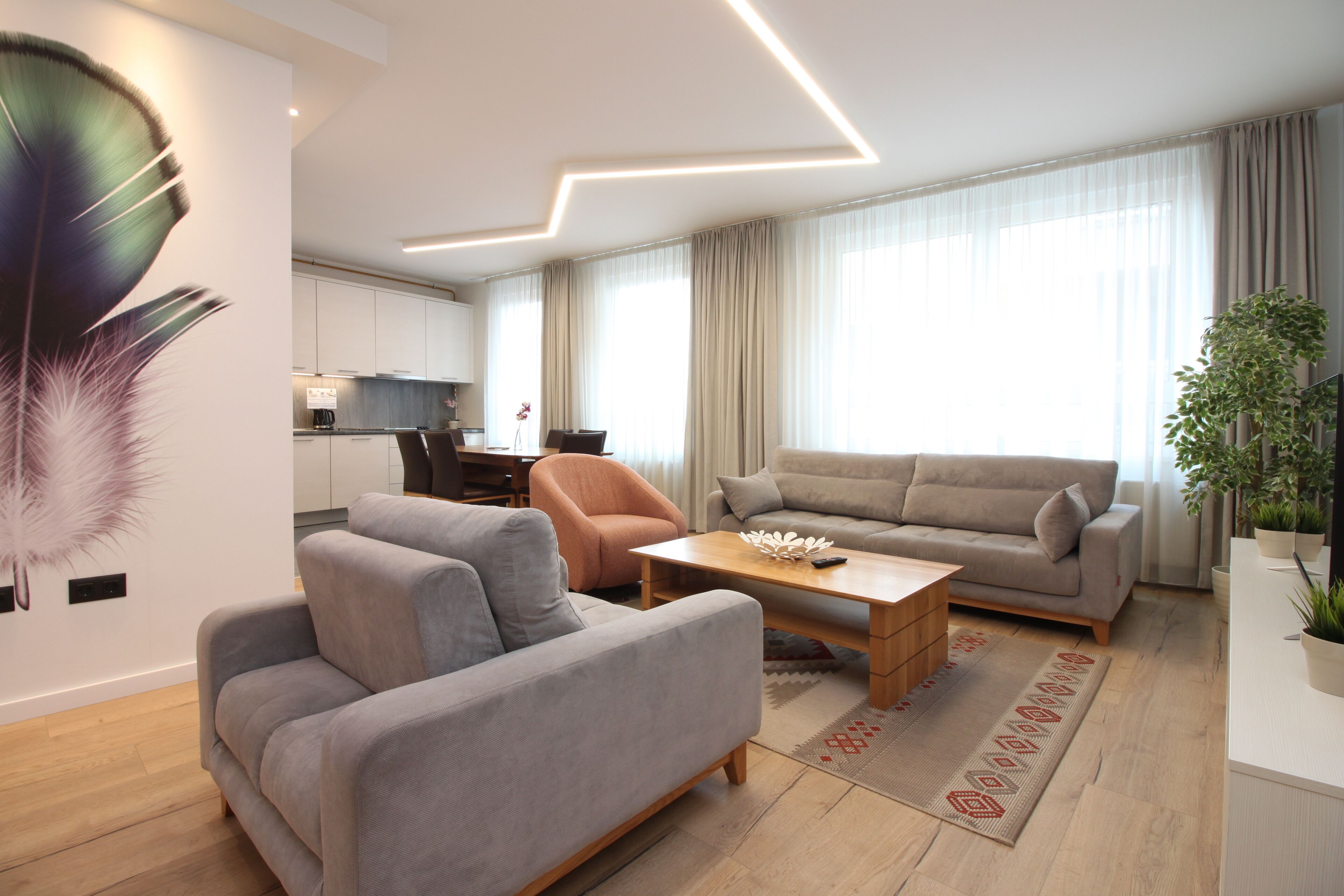 A modern living room with a grey sofa, a wooden coffee table, and a beige sofa. There is a dining area in the background with a kitchen visible.