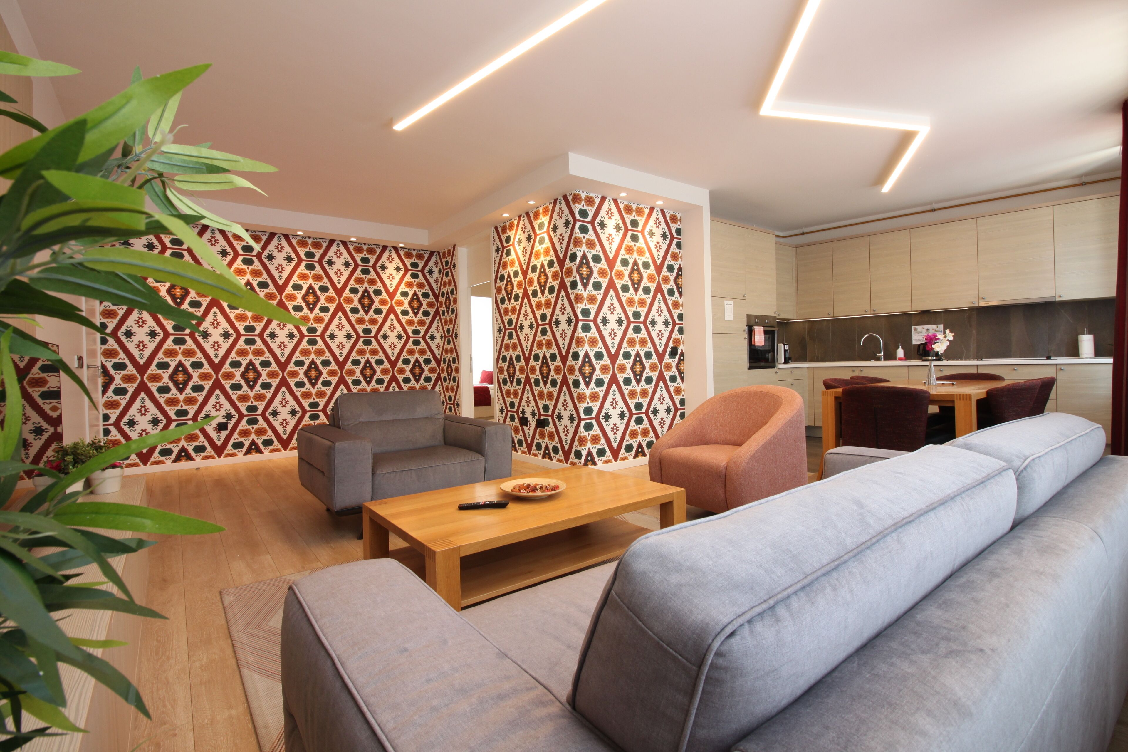 A modern living room with a sofa, armchairs, and a coffee table. There is a kitchen area with wooden cabinets and a dining table.