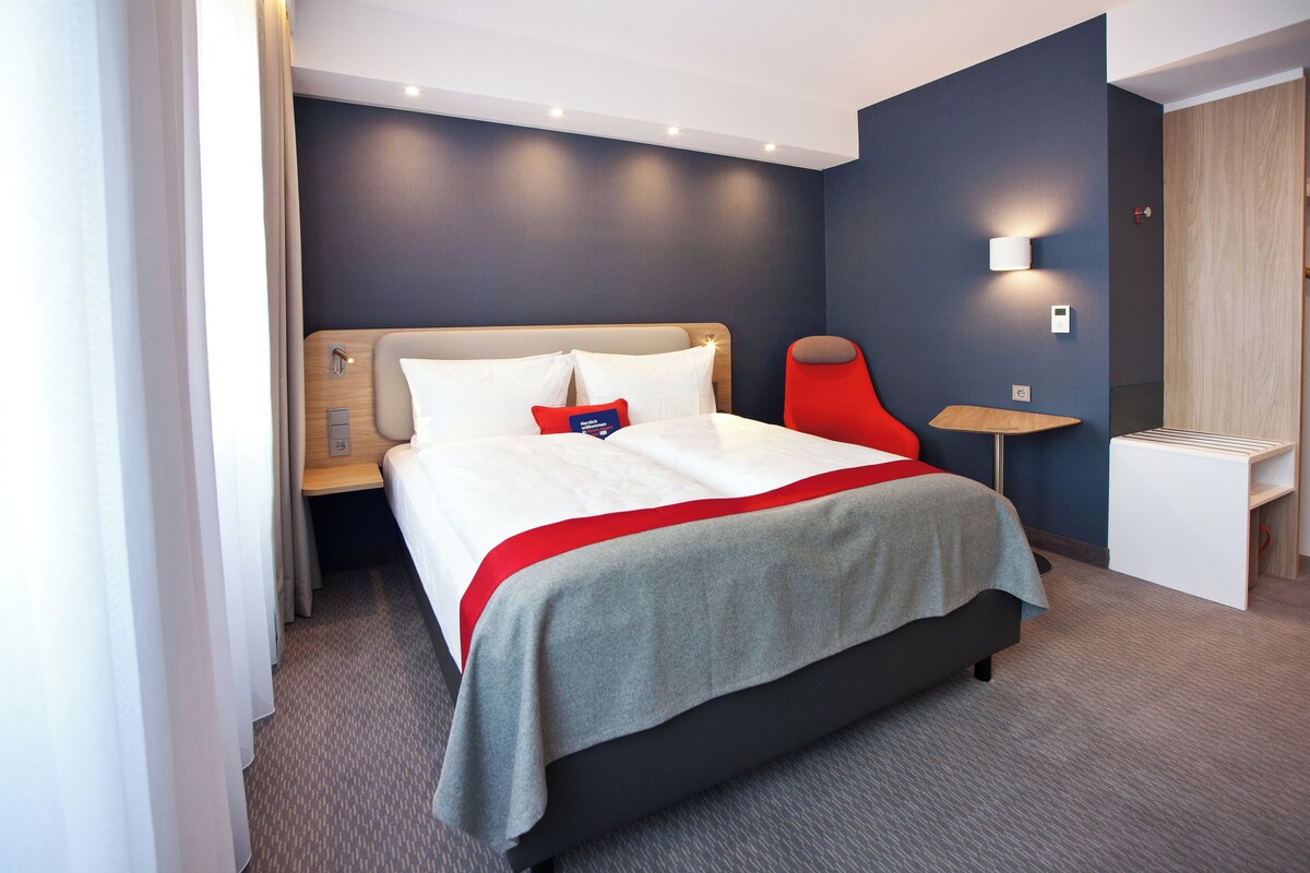 A modern hotel room with a bed, bedside table, and a small desk with a chair.