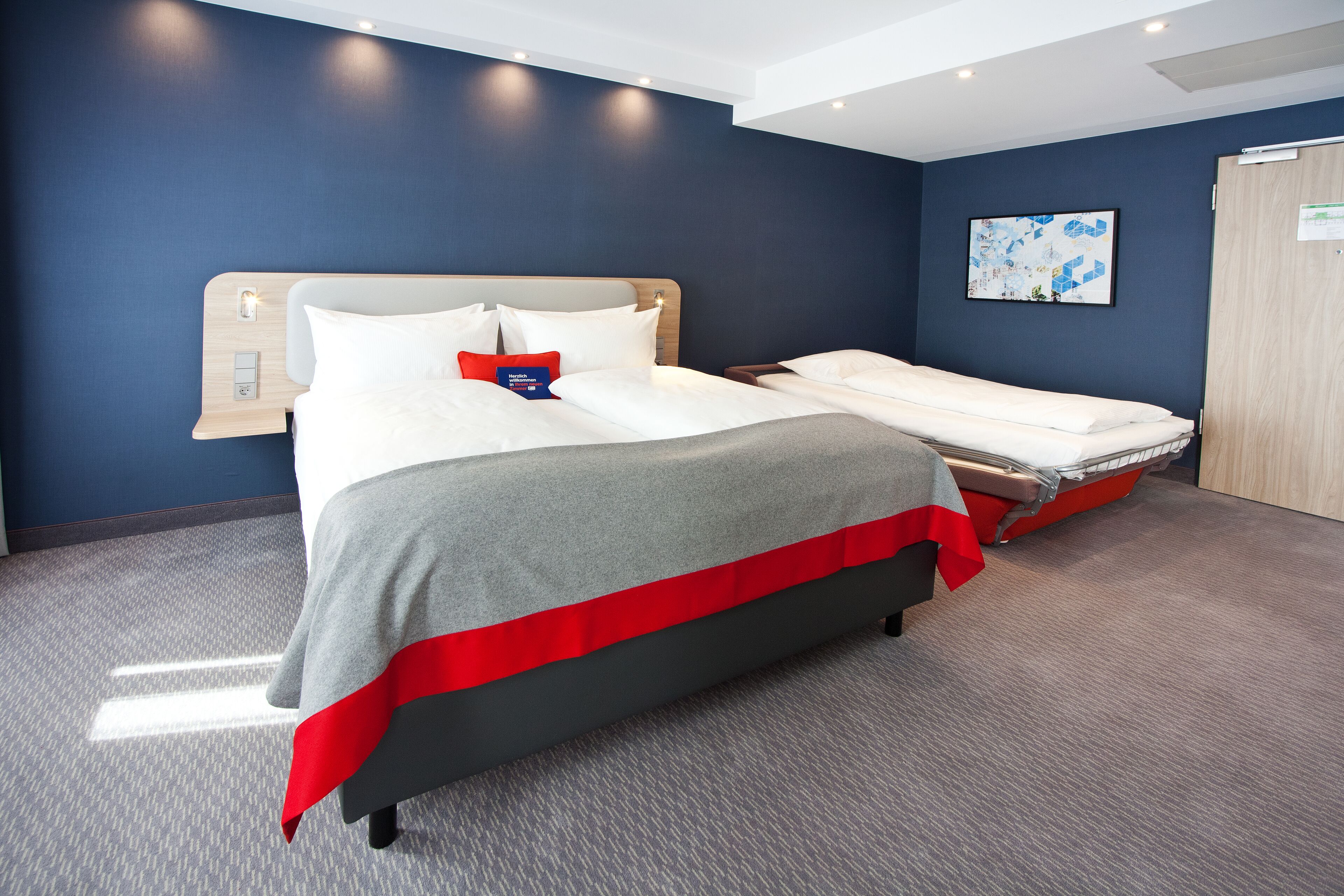 Una habitación de hotel con una cama grande, una cama más pequeña, una mesita de noche, un reloj en la pared y una puerta.