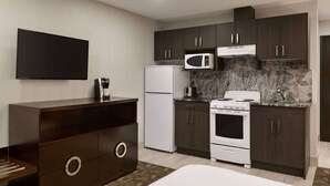 A compact kitchen with dark cabinets, a white refrigerator, a microwave, a stove, and a flat-screen TV. - Days Inn by Wyndham Wainwright (Wainwright)