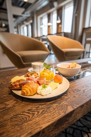 A breakfast plate with croissants, ham, cheese, and fruit on a wooden table in a modern hotel room with large windows and comfortable seating. - MASEVEN München Messe Dornach (Aschheim)