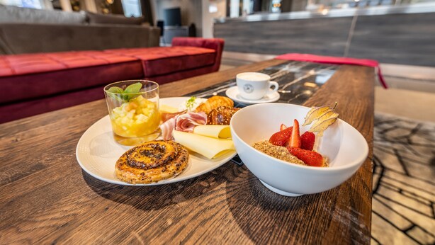 A breakfast setting with a plate of croissants, a bowl of cereal with strawberries, and a cup of coffee on a wooden table in a modern hotel lobby. - MASEVEN München Messe Dornach (Aschheim)