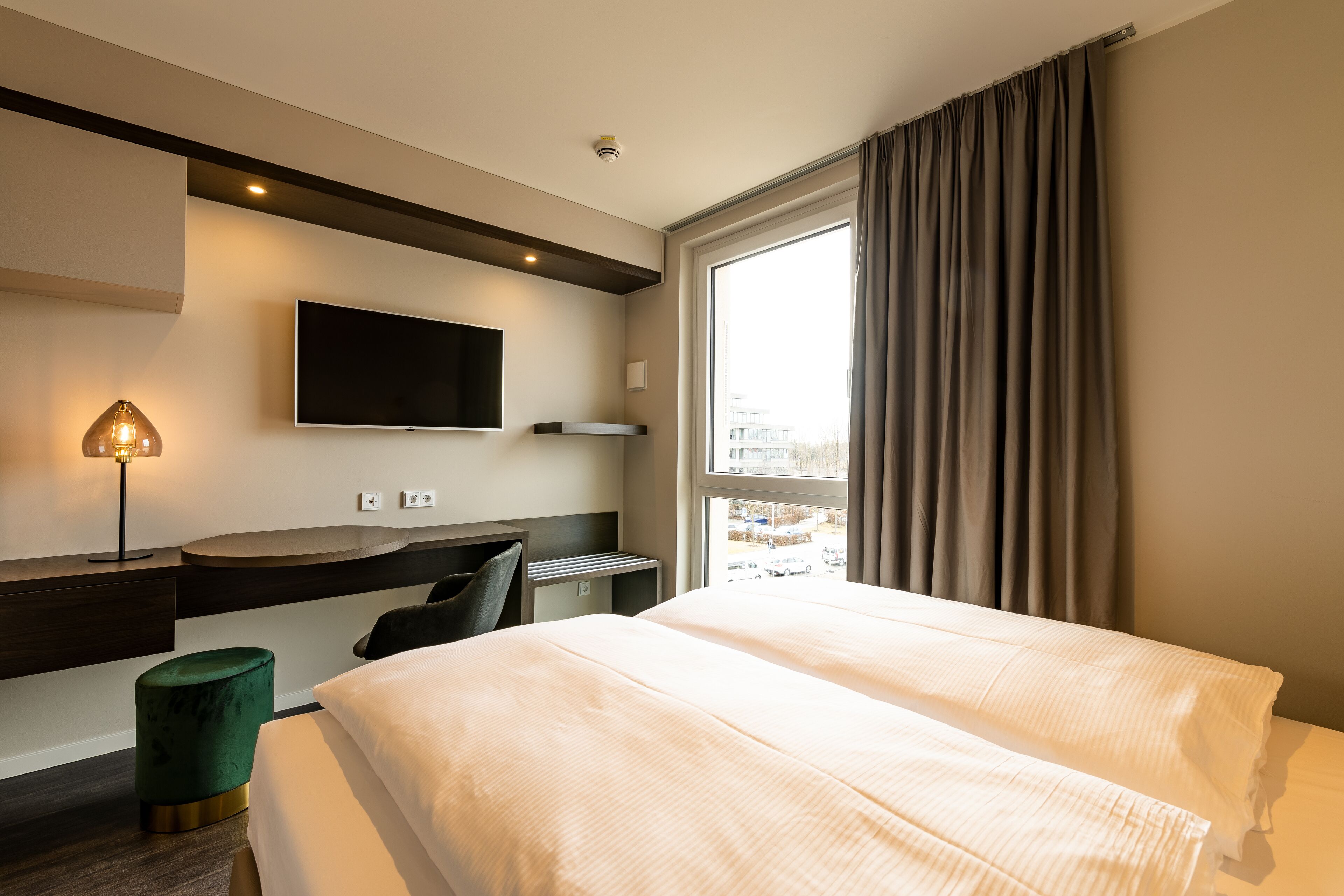 A modern hotel room with a large bed, a flat-screen TV mounted on the wall, a desk with a chair, and a window with curtains.