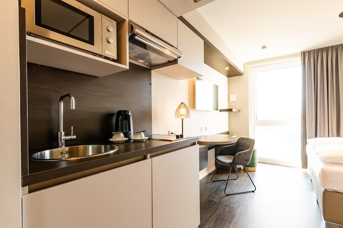 A compact hotel room with a kitchenette, a bed, a chair, and a large window.