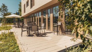 An outdoor patio with tables and chairs, a covered seating area, and a building with large windows. - MASEVEN München Messe Dornach (Aschheim)