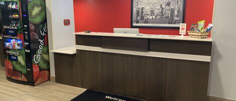 A reception area with a red wall, a framed welcome sign, a vending machine, and a Dell computer on the counter.