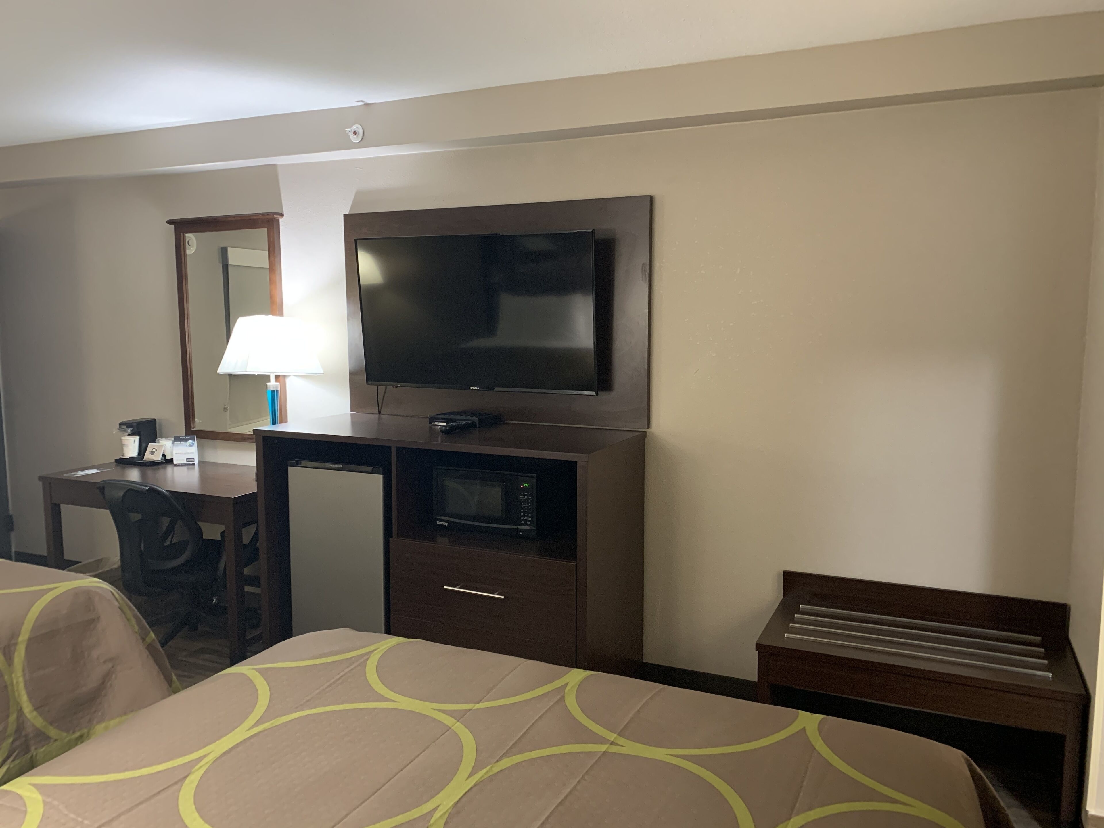 A hotel room with a bed, a desk with a lamp, a television, a microwave, and a small table.