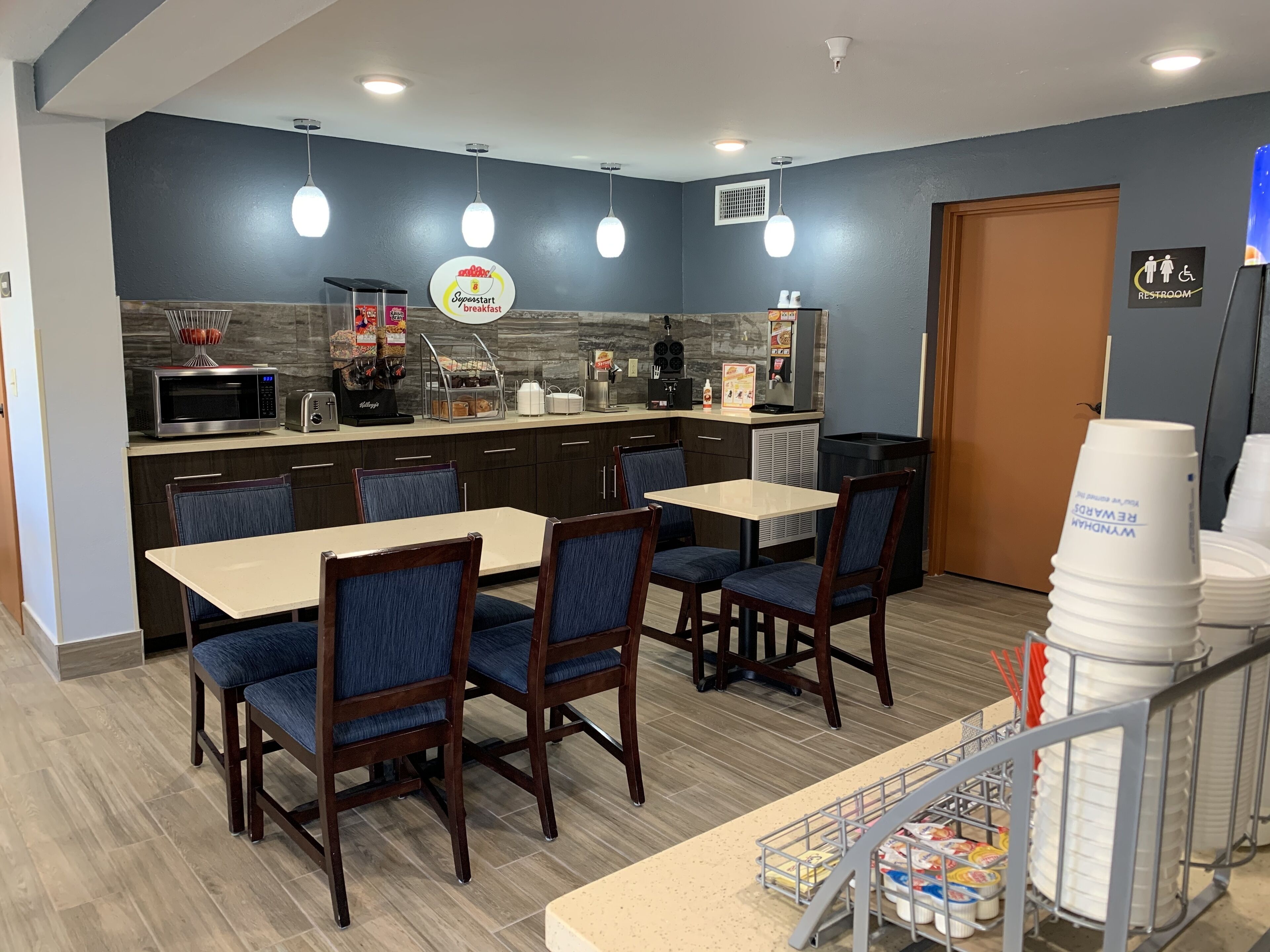 A dining area with a counter, microwave, and coffee machine. There are tables and chairs, and a sign for a restroom.