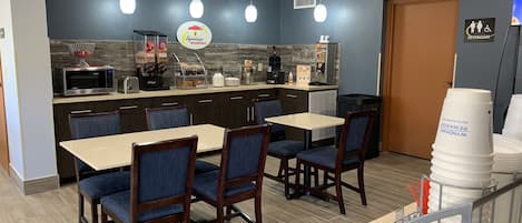 A dining area with a counter, microwave, and coffee machine. There are tables and chairs, and a sign for a restroom.