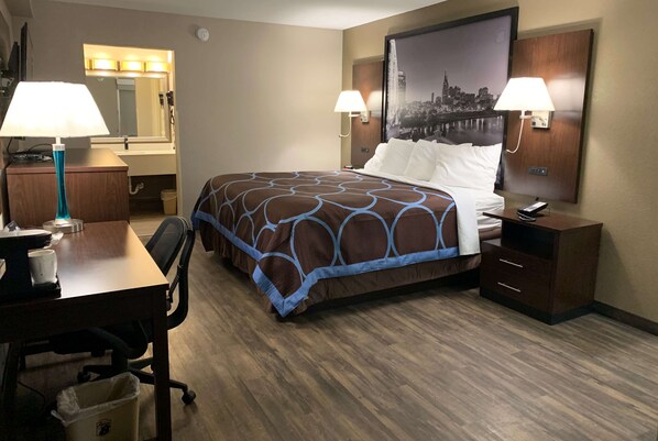 Quarto de hotel com cama, duas luminárias de cabeceira, uma escrivaninha com luminária, uma lixeira e uma TV fixada na parede.