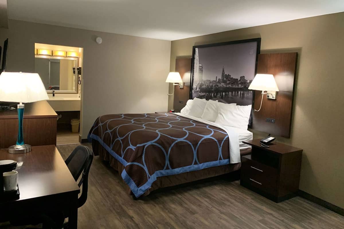 Une chambre d’hôtel avec un lit, deux lampes de chevet, un bureau avec une chaise, un miroir et un tableau représentant un paysage urbain accroché au mur.