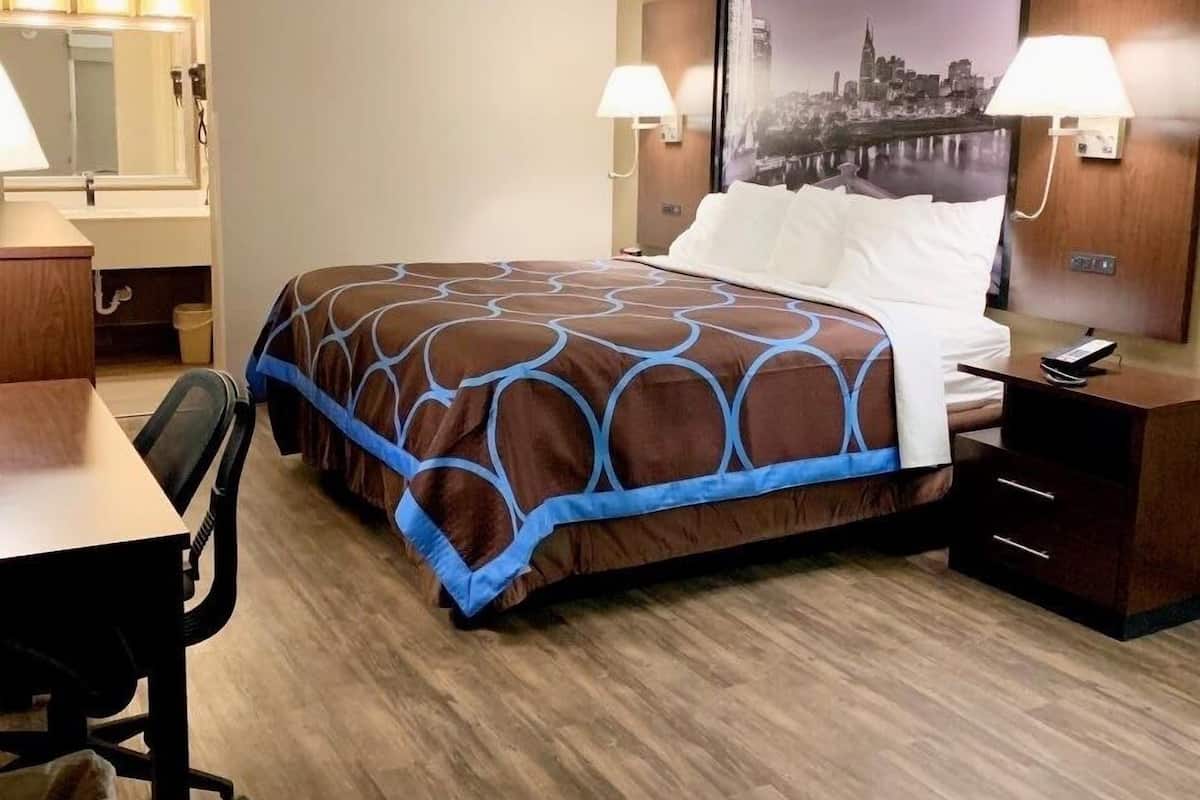 Une chambre d’hôtel avec un lit, un bureau, une chaise, une table de chevet, une lampe et un grand tableau représentant une vue sur la ville, accroché au mur.