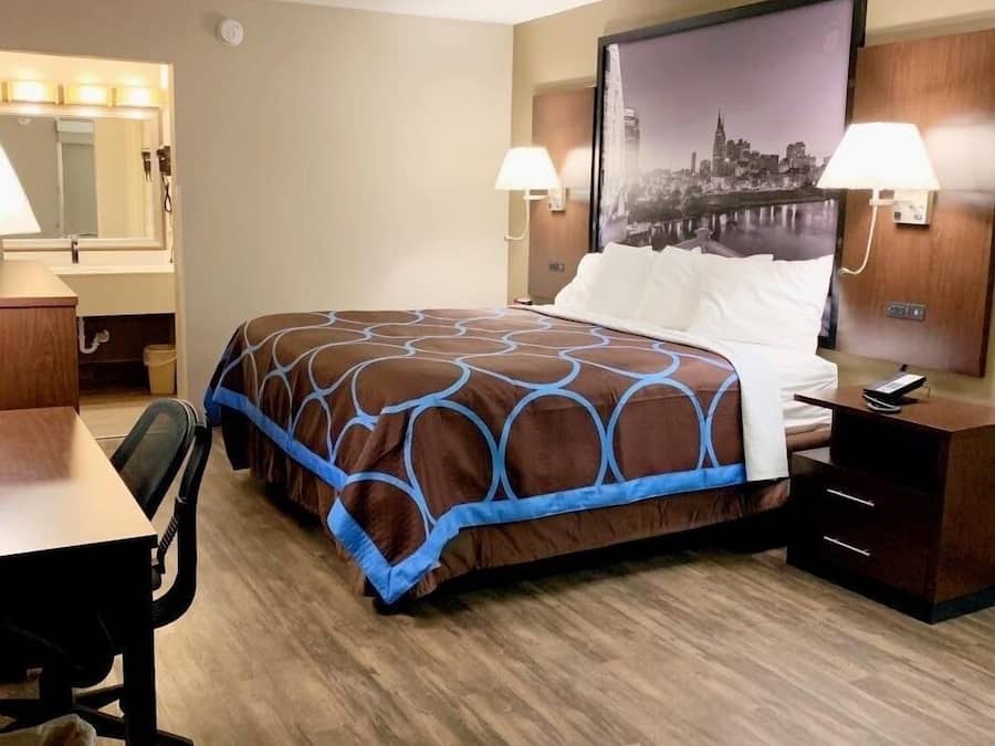 A hotel room with a bed, a desk, a chair, a nightstand, a lamp, and a large cityscape picture on the wall.