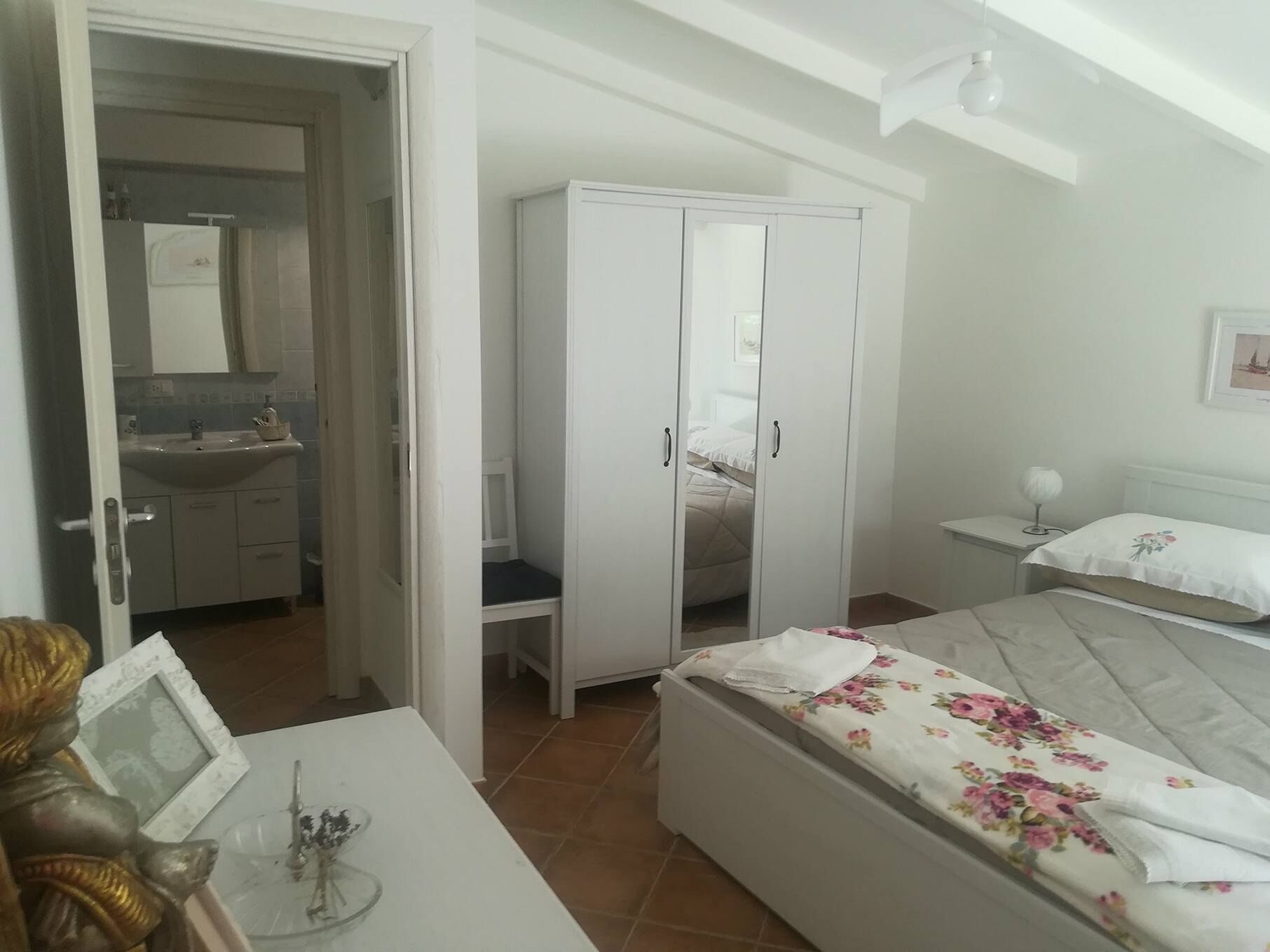 Une chambre à coucher avec un lit, une armoire, une table de chevet et une salle de bain visible à travers une porte ouverte.
