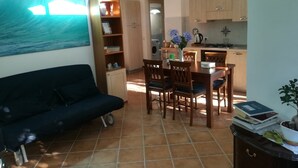 A living room with a dark blue sofa, a dining area with wooden chairs and a table, a kitchen with wooden cabinets, and a large painting on the wall. - L'Aranceto Sorrentino Residence (Meta)