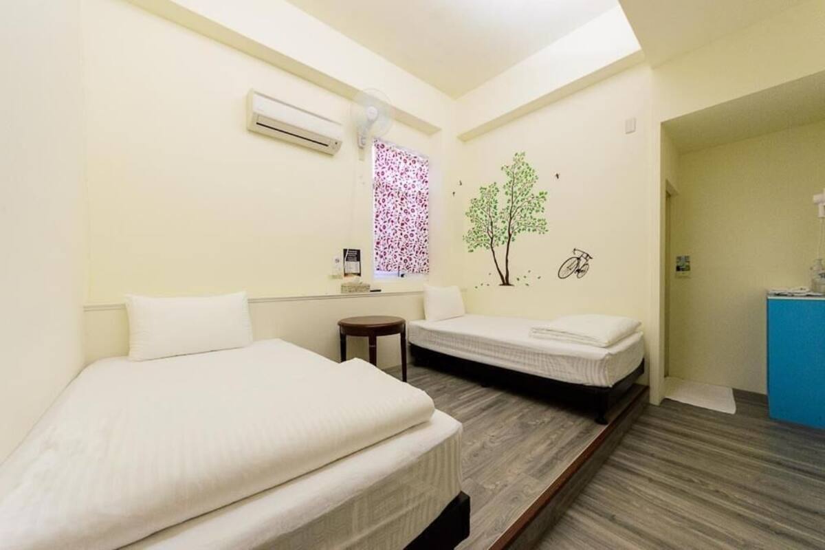 A hotel room with two beds, a small table, a window with a floral pattern, and a wall-mounted air conditioner.