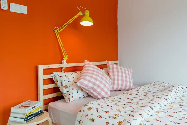 A bed with a floral-patterned bedspread, a wooden headboard, and two pillows with checkered patterns. A yellow desk lamp is on the wall beside the bed. - Eastern Court 22 by Sanguine (George Town)