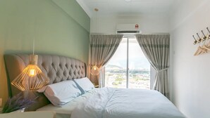 A bedroom with a tufted headboard, a bed with white bedding, a hanging light fixture, and a view of the city through large windows. - Eastern Court 22 by Sanguine (George Town)