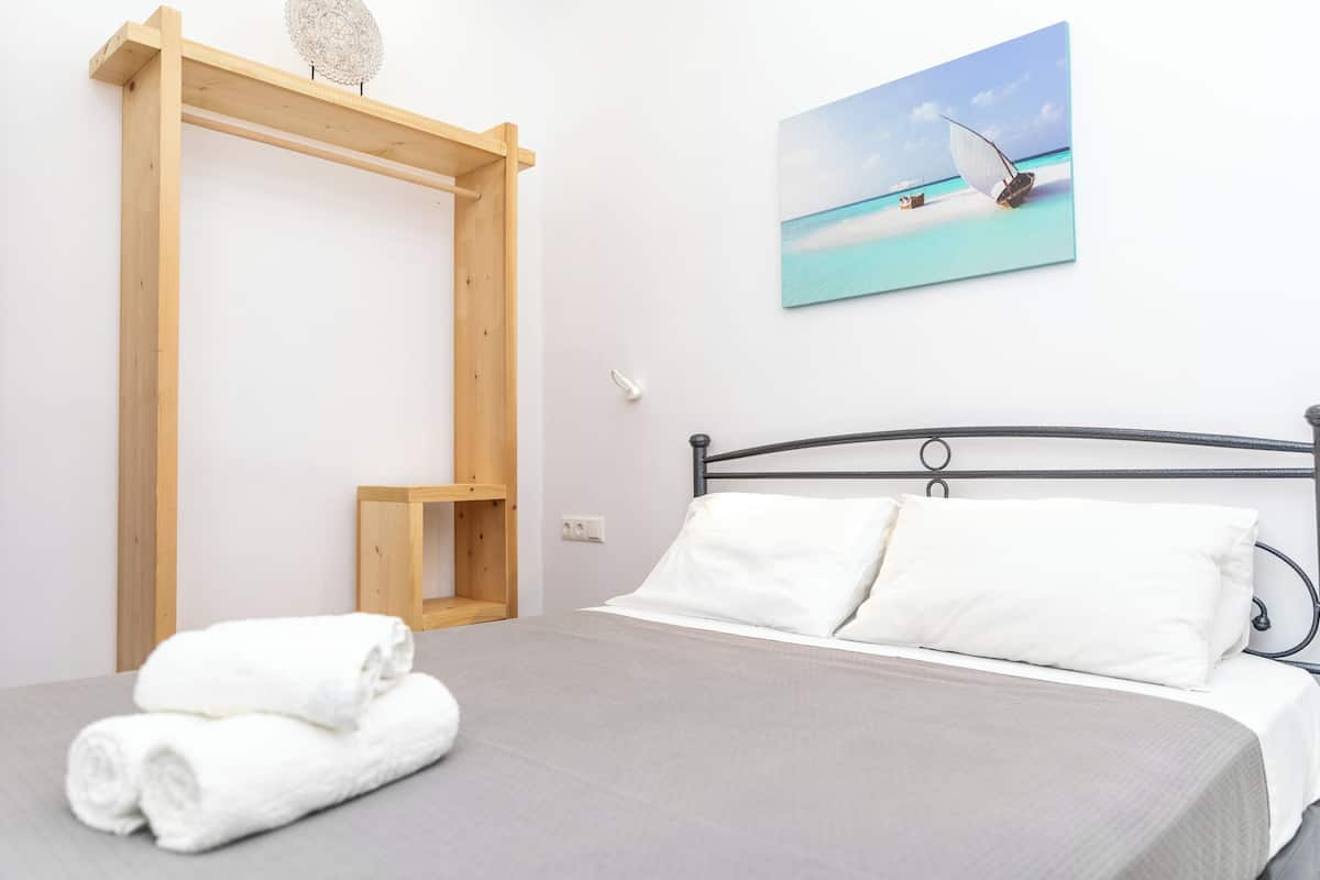A bedroom with a bed, white pillows, a grey bedspread, a wooden shelf, and a painting of a boat on water.
