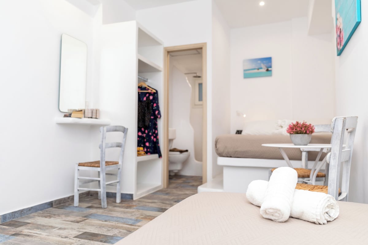 A modern bedroom with a bed, a chair, a small table with a plant, and a bathroom visible through an open door.