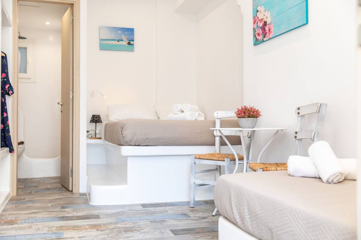 A modern bedroom with a bed, a small table with a chair, a window with a view of the sea, and a bathroom with a bathtub and toilet.