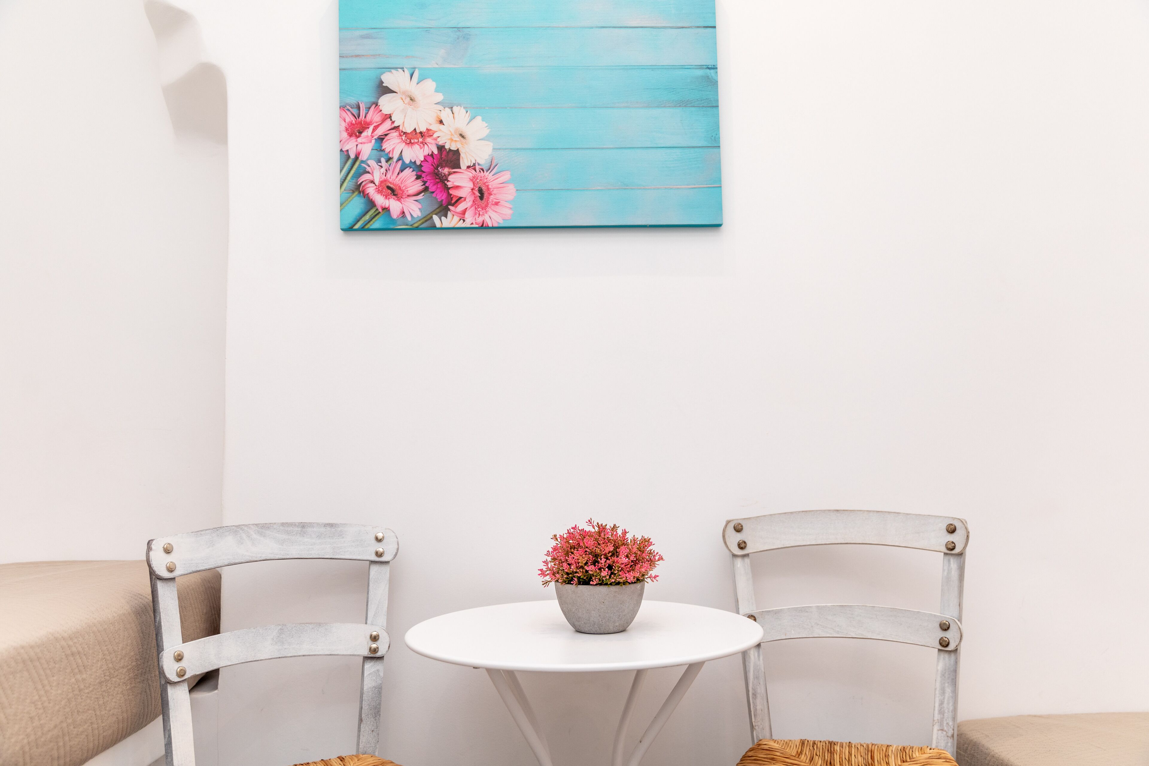 A room with a white table, two chairs, a painting of flowers, and a potted plant.