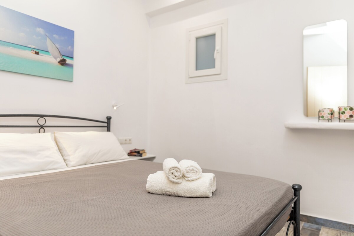 Una cama bien tendida con sábanas blancas y toallas enrolladas, una ventana pequeña con vistas, y un cuadro en la pared.