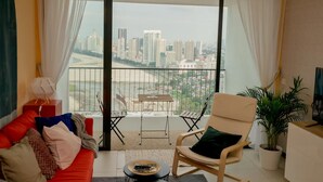 A living room with a red sofa, a white armchair, a small table, and a view of a cityscape through a balcony.