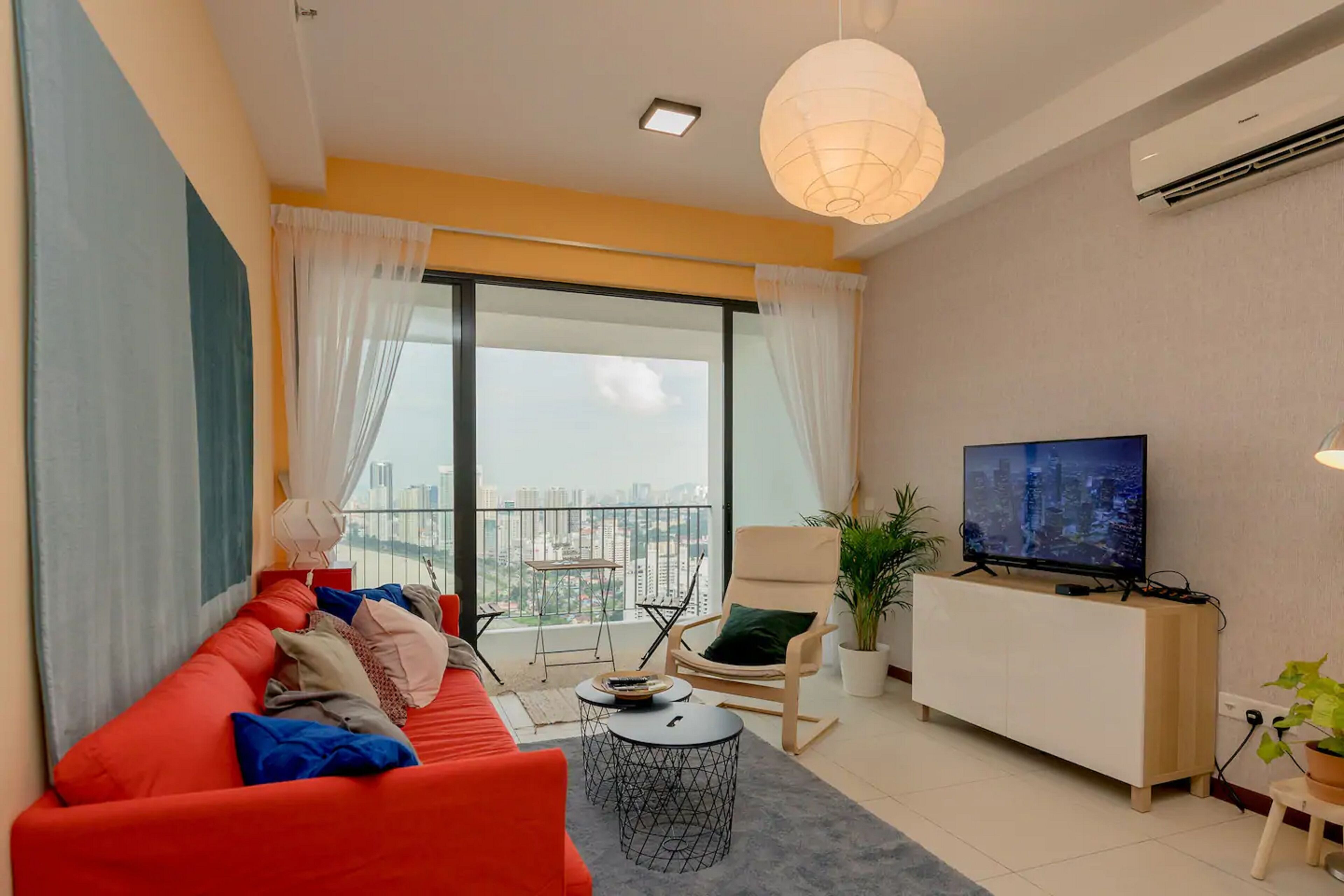 A modern living room with a red sofa, a TV, a balcony with a view of the city, and a potted plant.