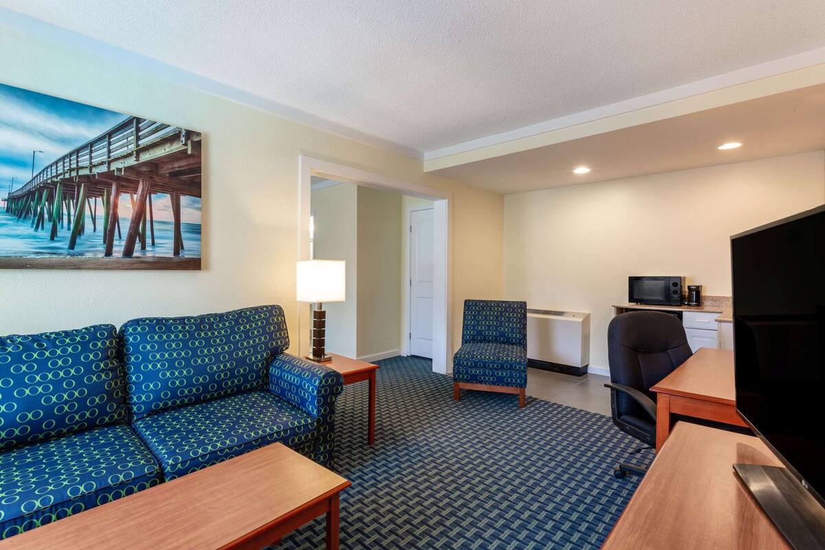 A hotel room with a sofa, armchair, TV, desk, and a painting of a pier.
