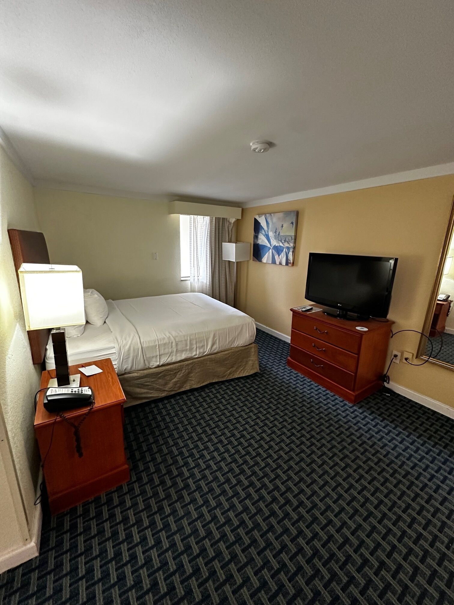 A hotel room with a bed, a television on a wooden dresser, a lamp, and a painting on the wall.