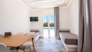Une chambre moderne avec une table en bois, deux chaises, un canapé, un téléviseur fixé au mur et une vue sur une piscine, visible à travers une porte vitrée.