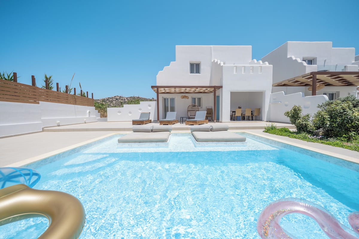 Een modern zwembadgedeelte met ligbedden en een pergola, omgeven door witte gebouwen in Mediterrane stijl.