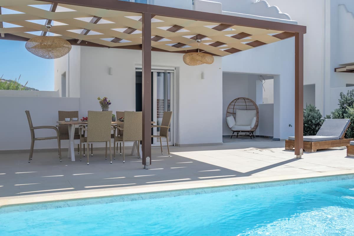 Een zwembadterras met een pergola, rieten meubels en een ligstoel.