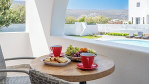 Une table ronde en bois, sur laquelle se trouvent une assiette de toasts, un saladier de fruits et deux tasses rouges, est placée sur un balcon donnant sur une piscine et un bâtiment blanc.