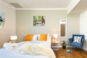 A hotel room with a bed, a chair, a painting, a mirror, and a nightstand with a lamp.
