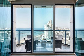 A balcony with two wicker chairs and a table, overlooking a cityscape and the sea.
