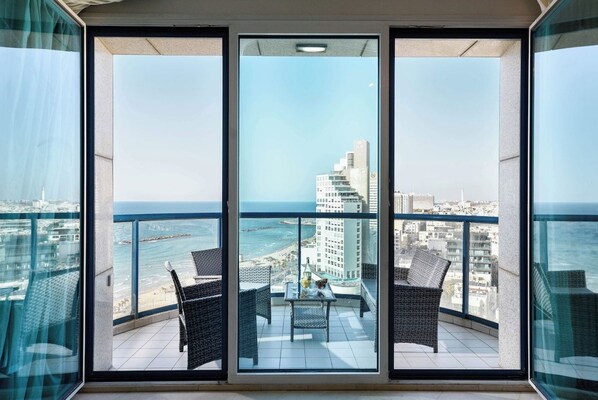 A balcony with two wicker chairs and a table, overlooking a cityscape and the sea.