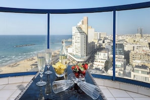 A table with champagne glasses, a bottle, and a fruit bowl with strawberries and pineapple, overlooking a beach and cityscape.