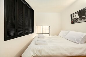 Ein Schlafzimmer mit einem Bett, weißen Bettwäsche, einem Fenster mit Jalousien und einem Schwarz-Weiß-Foto an der Wand.