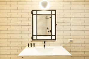 Ein Badezimmer mit einem weißen Waschbecken, einem Spiegel mit schwarzem Rahmen und einer sichtbaren Dusche im Spiegelbild.