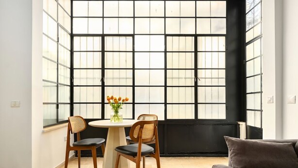A room with a large window, a round table with chairs, a sofa, and a vase with flowers. - Chic 1BR Apt Featuring Urban Design in the City (Tel Aviv-Yafo)
