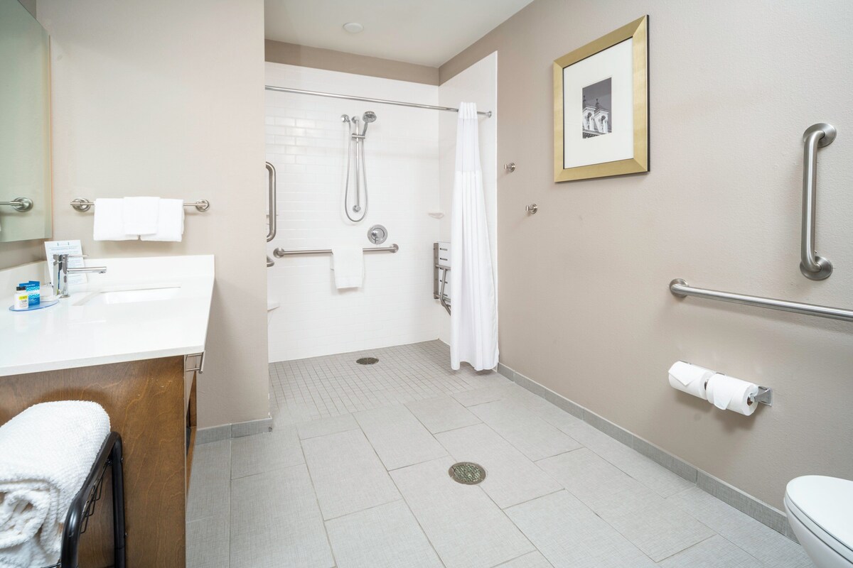 A bathroom with a white sink, a toilet, a shower with grab bars, and a framed picture on the wall.
