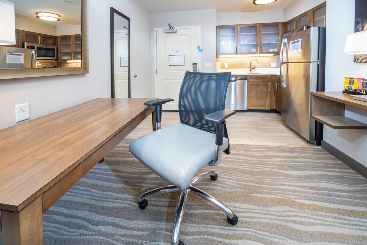 A conference room with a wooden table, a modern office chair, a kitchen area with stainless steel appliances, and a patterned carpet.
