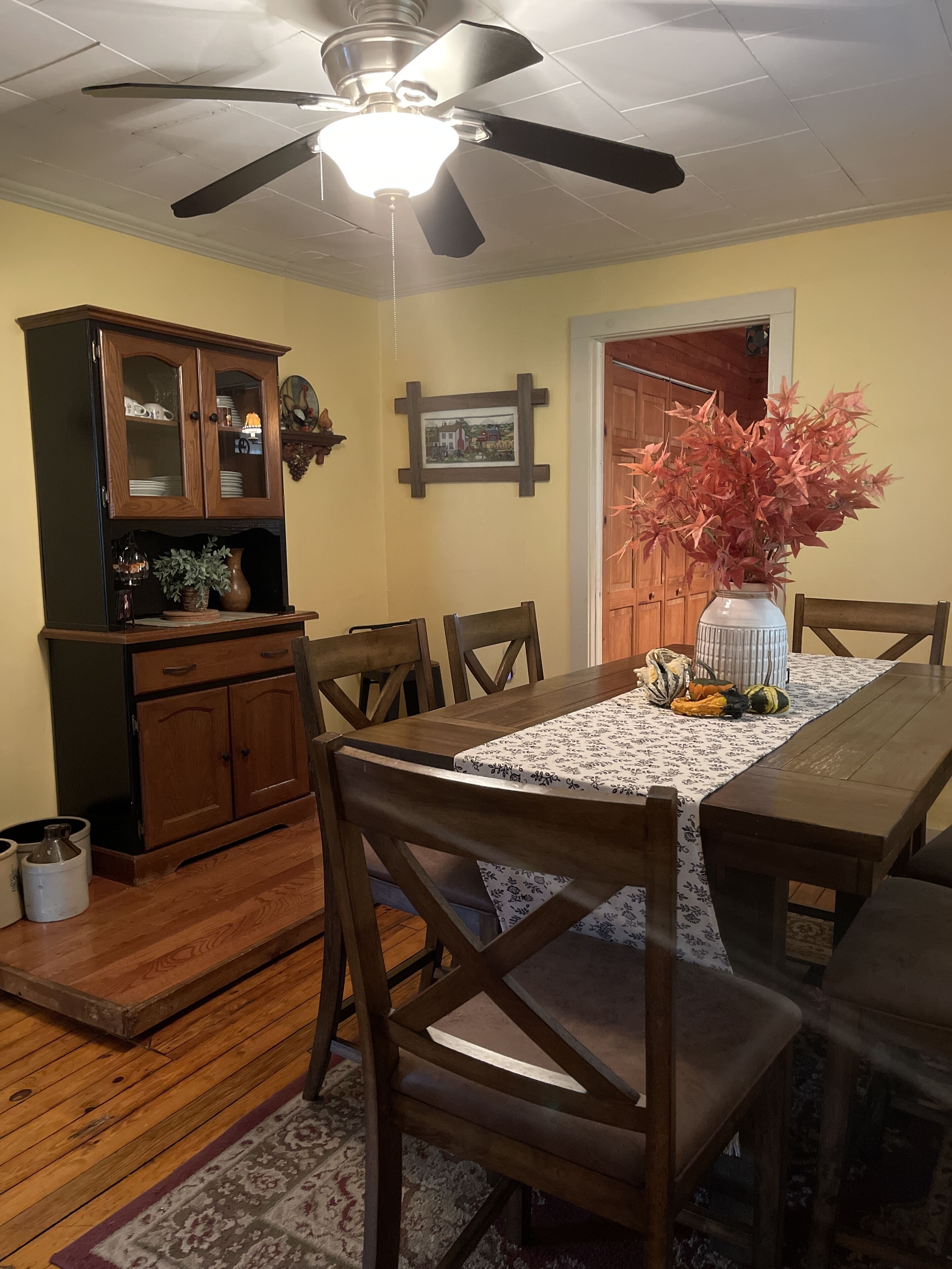 Un comedor con una mesa de madera, sillas, un ventilador de techo, un armario y un jarrón con flores.