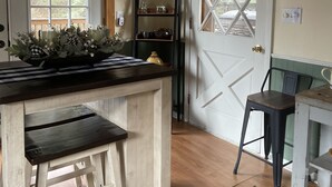 Una cocina con una mesa de madera, taburetes, un estante con adornos y una puerta con un diseño de ventana geométrico.