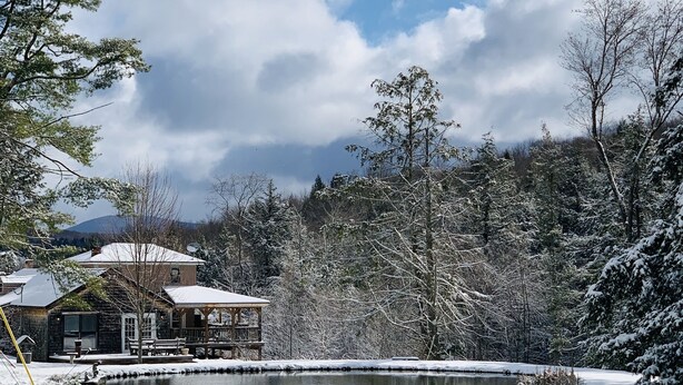 Paisaje nevado con un estanque, una estructura de madera y una casa al fondo.